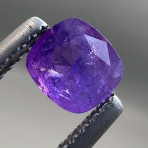 May include: A faceted, cushion-cut purple gemstone held by silver tweezers. The gem has a vibrant, translucent colour and a polished surface, reflecting light. The tweezers provide a contrasting metallic texture.
