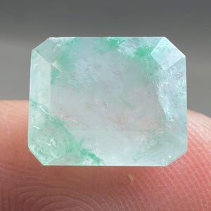 Op de afbeelding: Een rechthoekige edelsteen met een smaragdgroene slijpvorm. De steen is voornamelijk helder met lichtgroene strepen. De facetten zijn duidelijk gedefinieerd en de vorm is symmetrisch. De edelsteen wordt door een vinger vastgehouden.