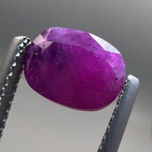May include: A faceted, oval-shaped, deep magenta gemstone is held by a pair of silver tweezers. The gem has a polished surface with multiple facets that reflect light. The background is a soft, neutral gray.