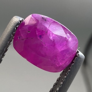 May include: A faceted, oval-shaped, deep pink gemstone held by silver tweezers. The gem has a polished surface with internal inclusions and a high degree of transparency. The gem is a vibrant shade of pink, with a slight purplish hue.