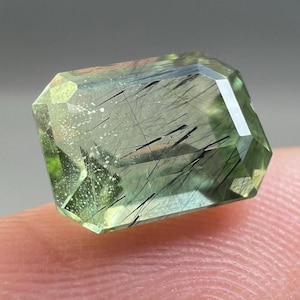 Puede incluir: Una gema rectangular facetada, de color verde pálido, con inclusiones negras. La gema se sostiene entre los dedos, mostrando sus superficies pulidas y patrones internos. Las facetas de la piedra capturan la luz, resaltando su claridad y color.