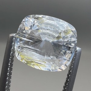 Puede incluir: Una gema transparente, tallada en cojín, sujeta por unas pinzas metálicas. La gema tiene inclusiones internas y un ligero tono amarillo-verdoso. Las facetas reflejan la luz, creando un efecto brillante.