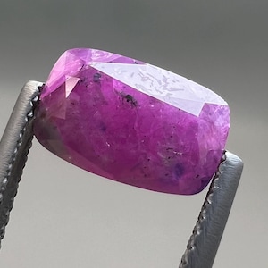 Puede incluir: Una gema facetada, de forma rectangular, de color rosa púrpura, sostenida por unas pinzas metálicas. La piedra pulida tiene múltiples facetas que reflejan la luz. El color de la piedra es un rosa vibrante y saturado con algunas inclusiones más oscuras.