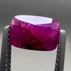 May include: A faceted, cushion-cut ruby gemstone held by a pair of metal tweezers. The ruby displays a deep, saturated red color with internal inclusions and a polished surface. The gemstone is rectangular with rounded corners.