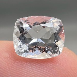 Puede incluir: Una piedra preciosa incolora y transparente, tallada en forma de cojín con una superficie facetada. La piedra presenta un aspecto claro similar al cristal con múltiples facetas geométricas que reflejan la luz y una pequeña inclusión.