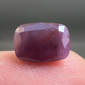 May include: A cushion-cut gemstone with a deep purple and magenta hue. The stone features a faceted surface that reflects light, showcasing a translucent quality with visible internal inclusions and a polished, smooth exterior finish.