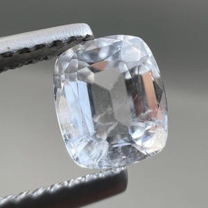 Può includere: Una gemma trasparente, taglio a cuscino, tenuta da una pinzetta. La gemma presenta molteplici sfaccettature e inclusioni interne, riflettendo la luce. La pinzetta è argentata e lo sfondo è grigio chiaro.