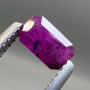 Op de afbeelding: Een diep magenta edelsteen, vastgehouden door een zilveren pincet, wordt getoond. De edelsteen is langwerpig en gefacetteerd, met een rechthoekige vorm. De facetten reflecteren licht, en kleine donkere insluitsels zijn zichtbaar.