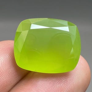 Pode incluir: Uma pedra preciosa facetada, de corte quadrado, com um tom verde limão vibrante. A superfície da gema reflete a luz, mostrando as suas múltiplas facetas e o acabamento polido. A gema é segurada numa mão, com um fundo neutro.