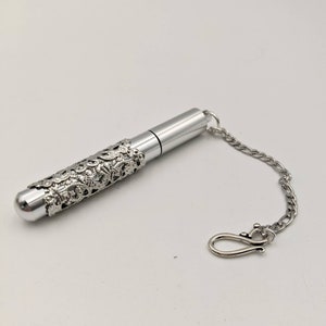 May include: A silver-colored, cylindrical container with a decorative filigree sleeve. The container has a rounded end and a chain with a clasp. The design is ornate and detailed, suggesting a vintage or antique style.