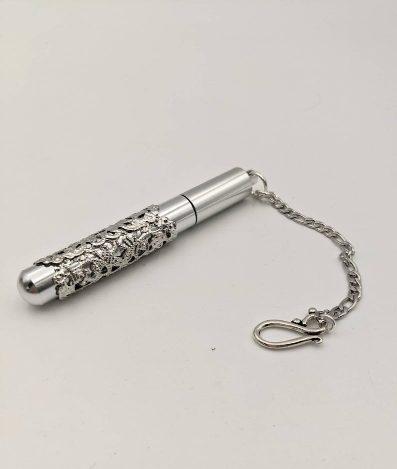 May include: A silver-colored, cylindrical container with a decorative filigree sleeve. The container has a rounded end and a chain with a clasp. The design is ornate and detailed, suggesting a vintage or antique style.
