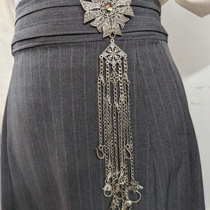 May include: A silver filigree brooch with a flower design and a long chain of dangling charms. The brooch is attached to a gray skirt.