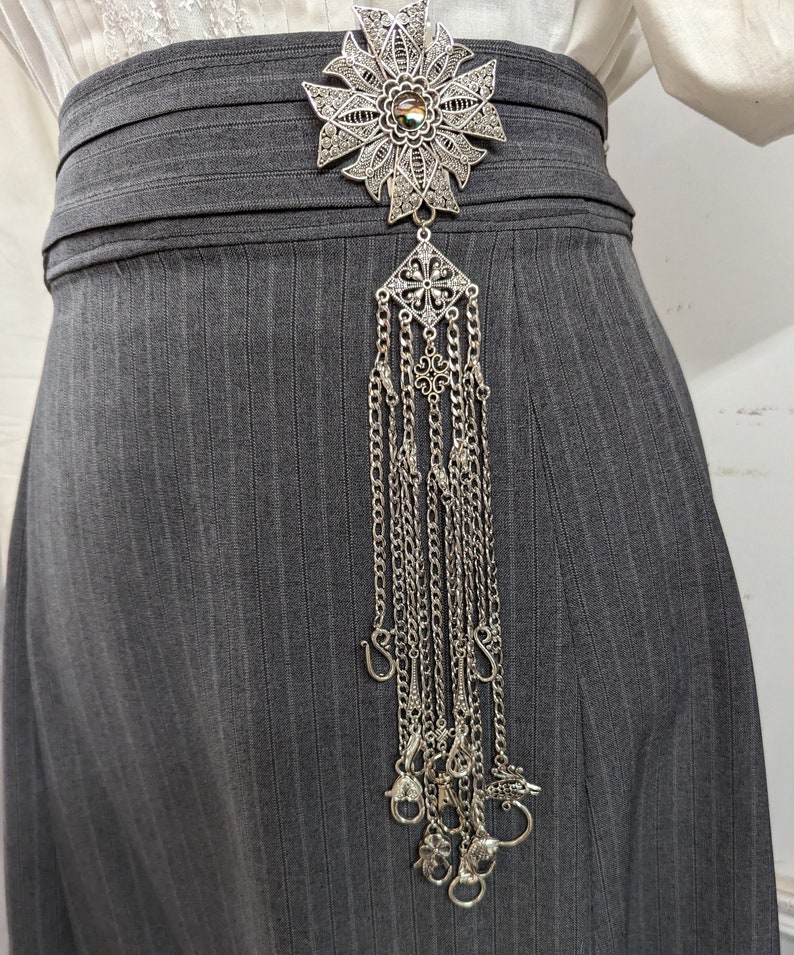 May include: A silver filigree brooch with a flower design and a long chain of dangling charms. The brooch is attached to a gray skirt.