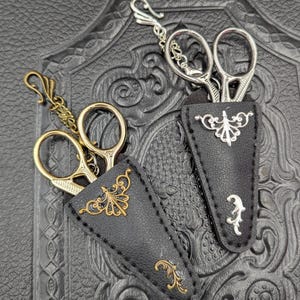 May include: Two pairs of decorative scissors with ornate handles and black leather sheaths. One pair has gold-colored accents, the other silver. The sheaths feature decorative metal embellishments. The scissors are designed for crafting or sewing.