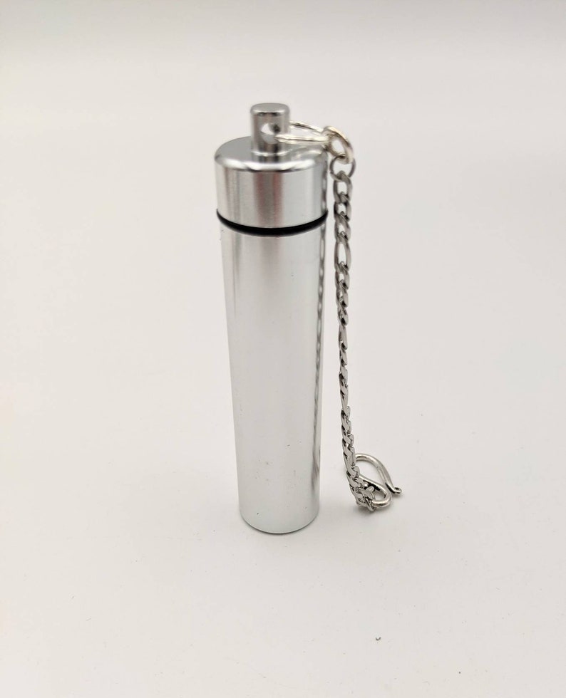 May include: A silver-colored, cylindrical urn with a screw-on top and a small chain. The urn is made of metal and has a polished finish. The chain is attached to the top and has a clasp. The urn is designed to hold a small amount of cremated remains.