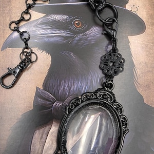 May include: Black metal pocket watch with an oval glass face and ornate frame, suspended from a chain with a lobster clasp. The watch hangs in front of a raven illustration, creating a gothic aesthetic.