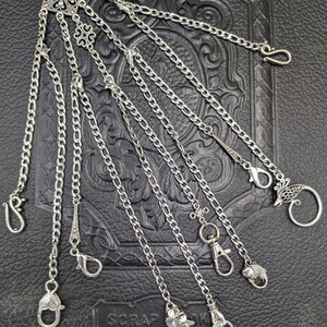 May include: A silver chain with multiple charms and attachments. The chain is made of multiple strands of silver chain and has a variety of charms, including a flower, a bird, a fish, and a heart. The chain is hanging on a dark background.