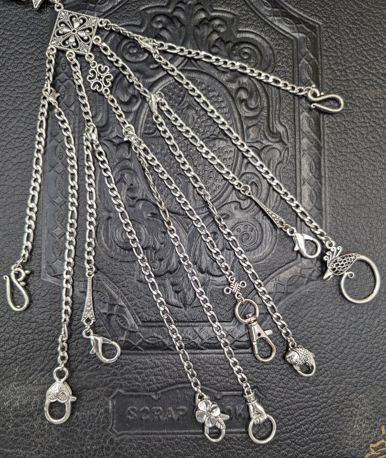May include: A silver chain with multiple charms and attachments. The chain is made of multiple strands of silver chain and has a variety of charms, including a flower, a bird, a fish, and a heart. The chain is hanging on a dark background.
