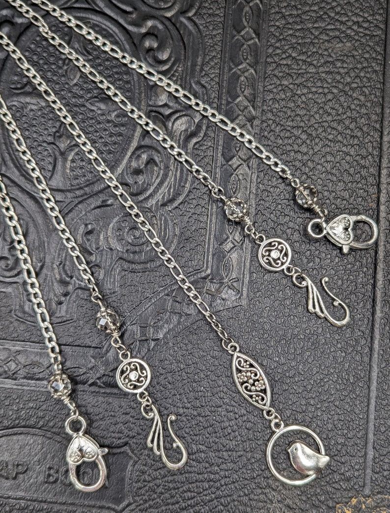 May include: Four silver chains with different decorative charms, including a bird, a flower, and a leaf. The chains are displayed on a textured black surface.