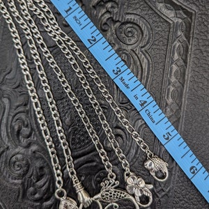 May include: Four silver chains with different sized links and decorative silver charms, including a hand, a bird, a flower, and a fish. The chains are laid out on a black leather surface with a blue measuring tape.