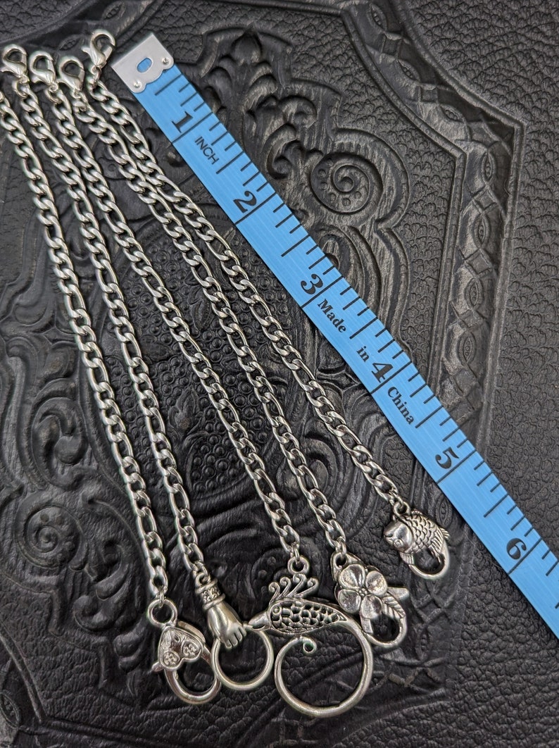 May include: Four silver chains with different sized links and decorative silver charms, including a hand, a bird, a flower, and a fish. The chains are laid out on a black leather surface with a blue measuring tape.