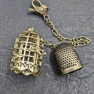 May include: A vintage-style charm featuring a bird cage, thimble, and bird-shaped clasp. The cage and thimble are gold-toned metal, connected by a chain. The bird clasp adds a decorative touch to this unique accessory.