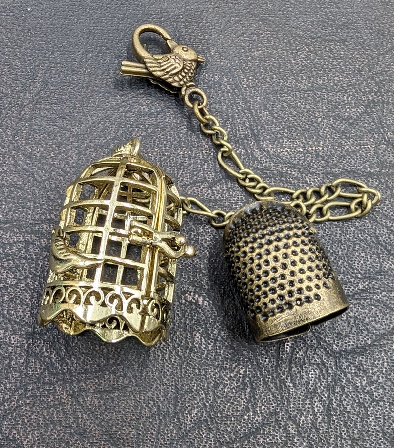 May include: A vintage-style charm featuring a bird cage, thimble, and bird-shaped clasp. The cage and thimble are gold-toned metal, connected by a chain. The bird clasp adds a decorative touch to this unique accessory.
