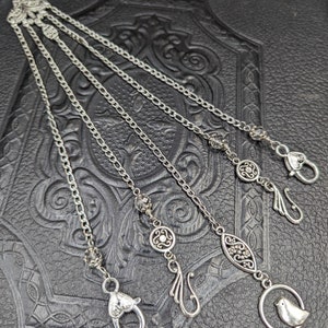 May include: A silver chain necklace with five charms, including a bird, a swirl, a circle, an oval, and a hook. The charms are attached to the chain with small crystal beads.