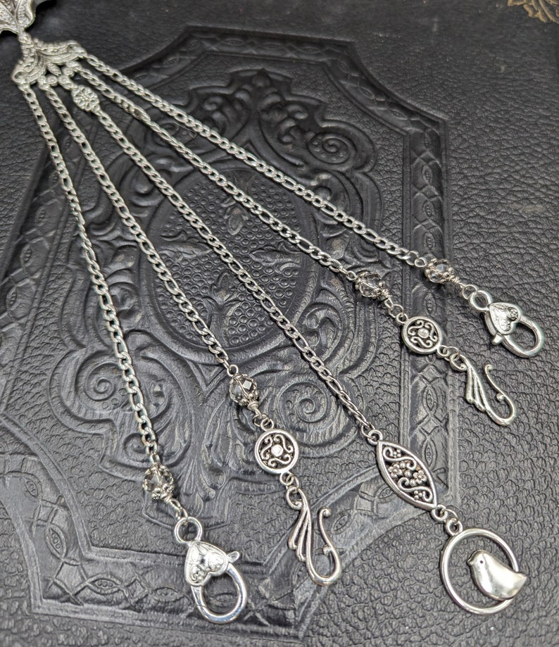 May include: A silver chain necklace with five charms, including a bird, a swirl, a circle, an oval, and a hook. The charms are attached to the chain with small crystal beads.