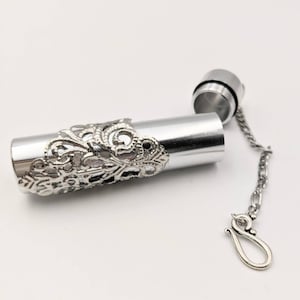 May include: A silver-colored, cylindrical container with an ornate, filigree design around the middle. The top is unscrewed, revealing the interior. A small chain with a hook is attached to the top.