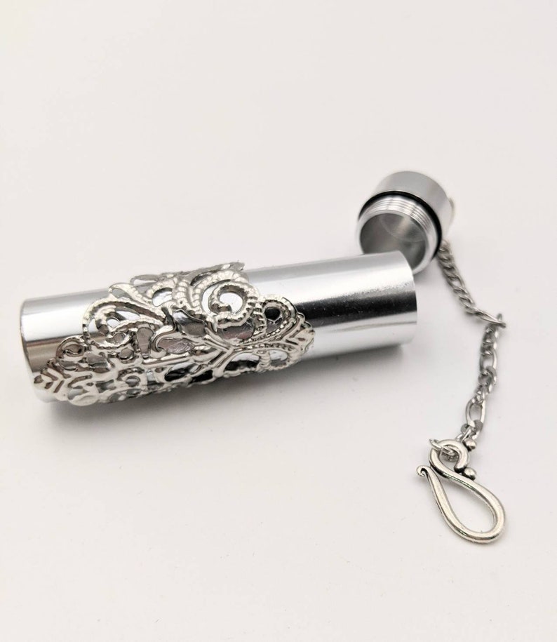 May include: A silver-colored, cylindrical container with an ornate, filigree design around the middle. The top is unscrewed, revealing the interior. A small chain with a hook is attached to the top.