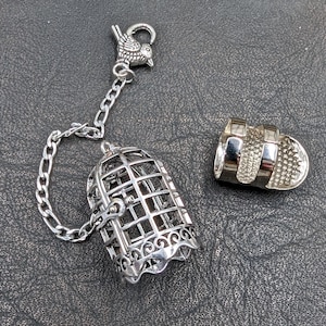 May include: A silver-colored tea infuser shaped like a birdcage, with a matching bird charm and chain. The infuser is open, with a separate lid. The birdcage has a decorative, scalloped base.