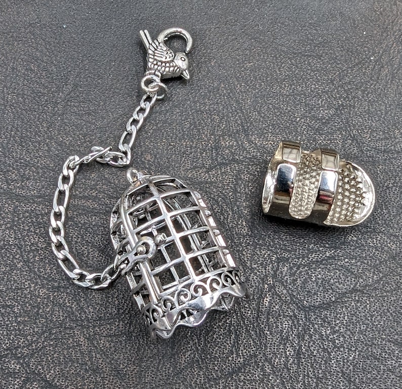 May include: A silver-colored tea infuser shaped like a birdcage, with a matching bird charm and chain. The infuser is open, with a separate lid. The birdcage has a decorative, scalloped base.
