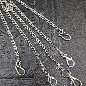 May include: A silver chain with multiple hooks and decorative accents. The chain is on a black leather background.
