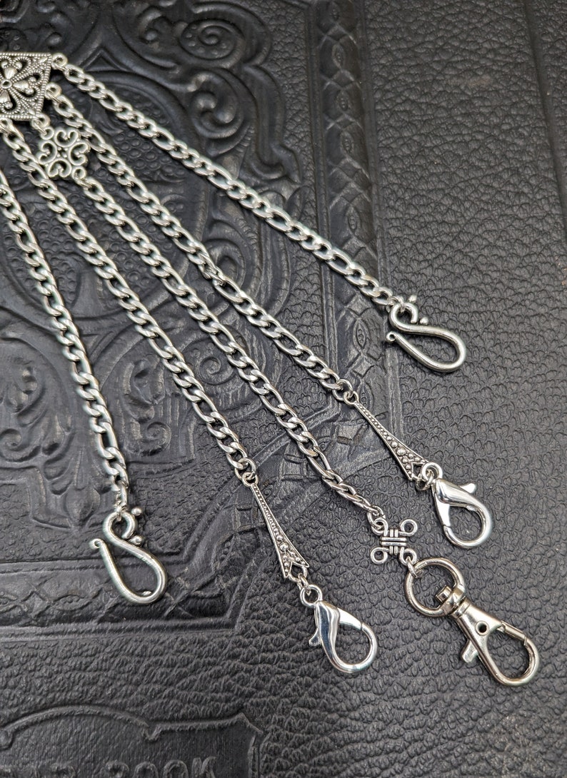 May include: A silver chain with multiple hooks and decorative accents. The chain is on a black leather background.