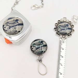 May include: A silver retractable tape measure with a black and white loon design. The tape measure has a silver chain attached to it. There is also a silver ruler with a black and white loon design and a silver clasp.