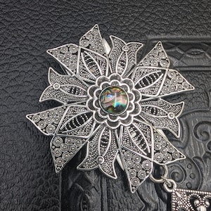 May include: A silver filigree flower brooch with a round iridescent abalone shell in the center. The brooch has a dangling pendant with a geometric design.