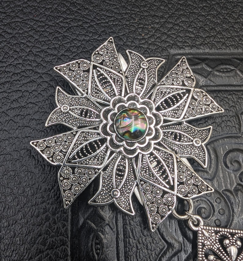 May include: A silver filigree flower brooch with a round iridescent abalone shell in the center. The brooch has a dangling pendant with a geometric design.