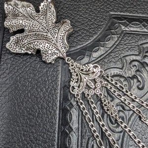 May include: Silver leaf bookmark with a detailed floral design and a chain tassel. The bookmark is on a black textured surface.