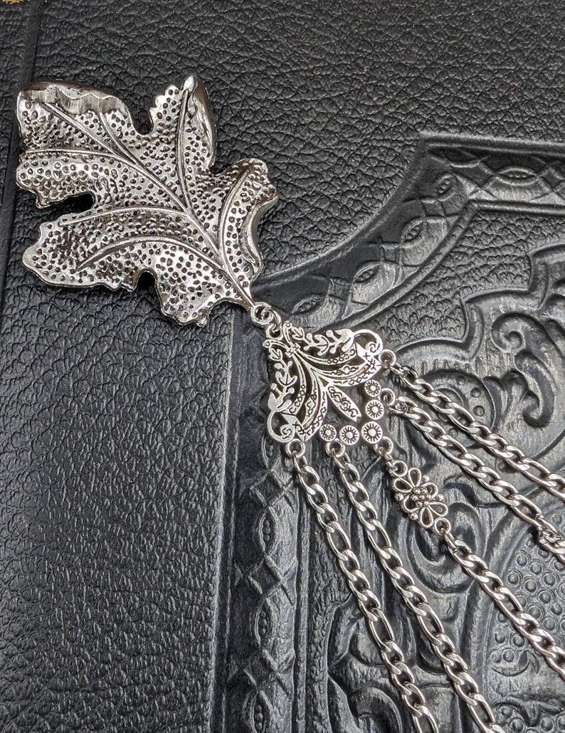 May include: Silver leaf bookmark with a detailed floral design and a chain tassel. The bookmark is on a black textured surface.