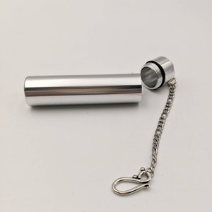 May include: A silver-colored, cylindrical container with a screw-on cap, connected by a chain to a decorative clasp. The container is open, revealing the threaded interior. The clasp has a decorative design.