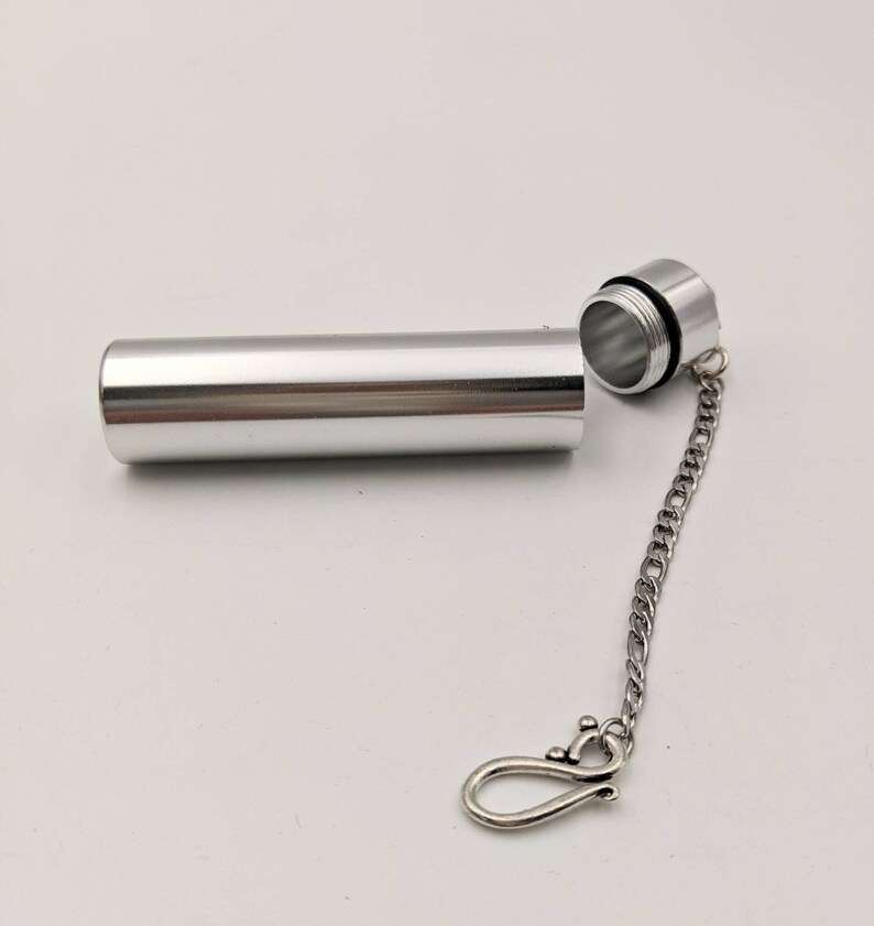 May include: A silver-colored, cylindrical container with a screw-on cap, connected by a chain to a decorative clasp. The container is open, revealing the threaded interior. The clasp has a decorative design.