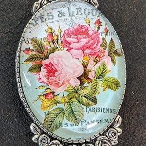 Può includere: Un ciondolo d'argento con un cabochon di vetro che presenta un design floreale vintage con rose rosa e foglie verdi. Il testo "Parisienne" e "Arts de Pansem" è visibile sul ciondolo.
