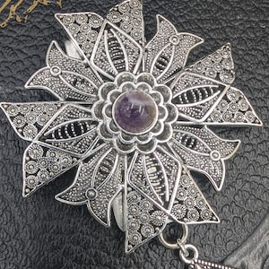 May include: A silver filigree pendant with a purple amethyst gemstone in the center. The pendant is in the shape of a flower with intricate details.