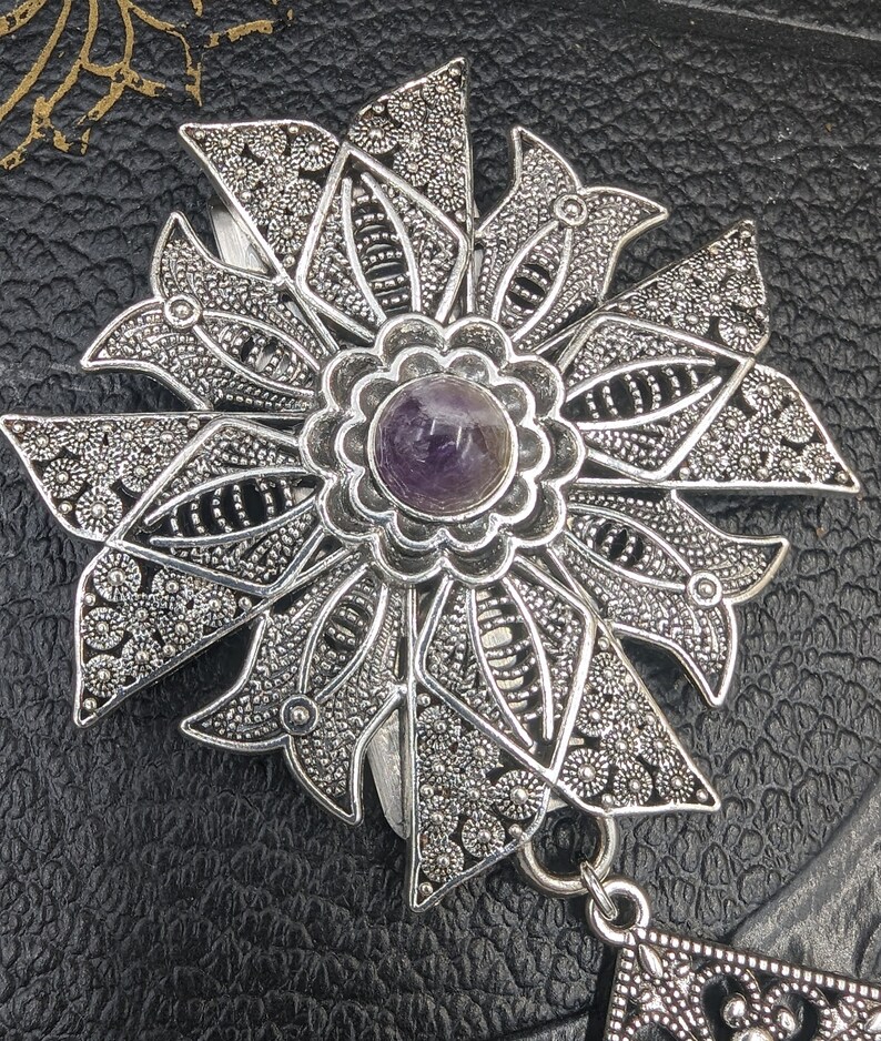 May include: A silver filigree pendant with a purple amethyst gemstone in the center. The pendant is in the shape of a flower with intricate details.