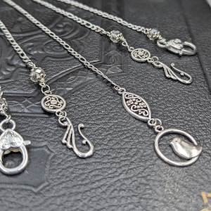 May include: Five silver chains with decorative charms, including a bird, a floral design, and a swirl. The chains are on a black leather surface.