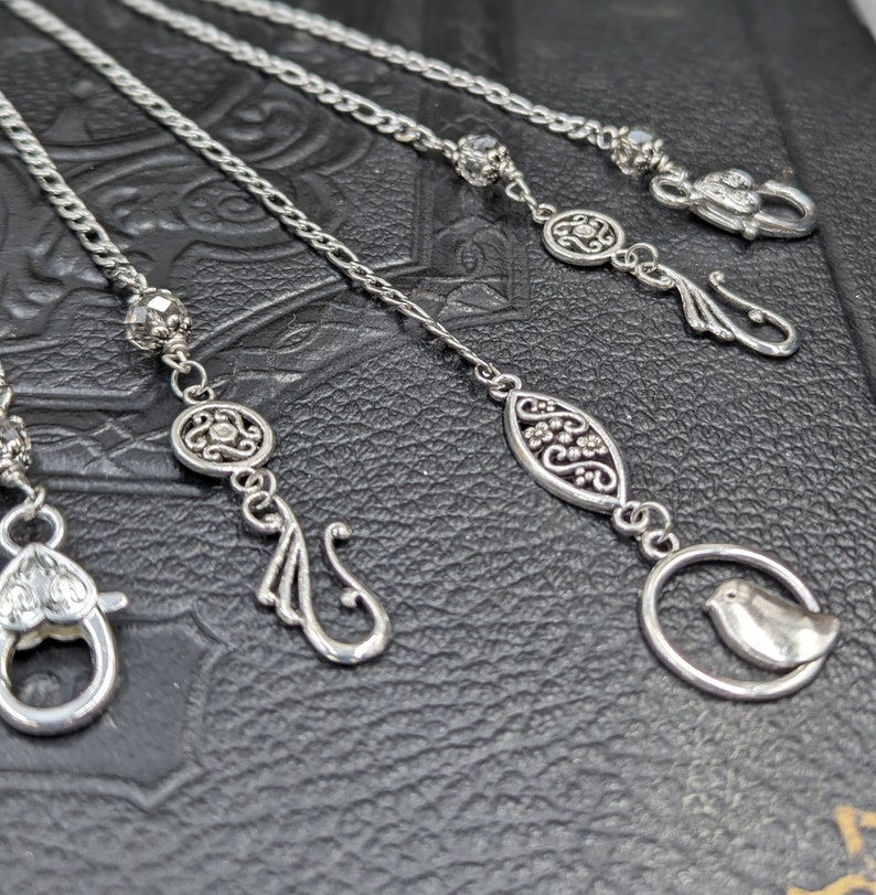 May include: Five silver chains with decorative charms, including a bird, a floral design, and a swirl. The chains are on a black leather surface.