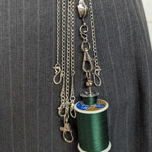 Puede incluir: Una cadena de plata con múltiples cierres y un carrete de hilo verde oscuro colgando de ella.