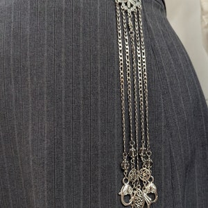 May include: A silver leaf-shaped brooch with a chain and decorative charms hanging from it. The brooch is attached to a gray pinstripe fabric.