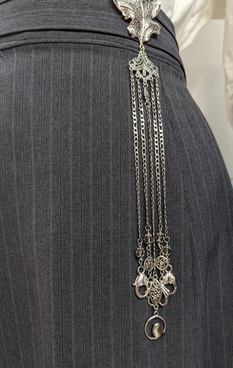 May include: A silver leaf-shaped brooch with a chain and decorative charms hanging from it. The brooch is attached to a gray pinstripe fabric.